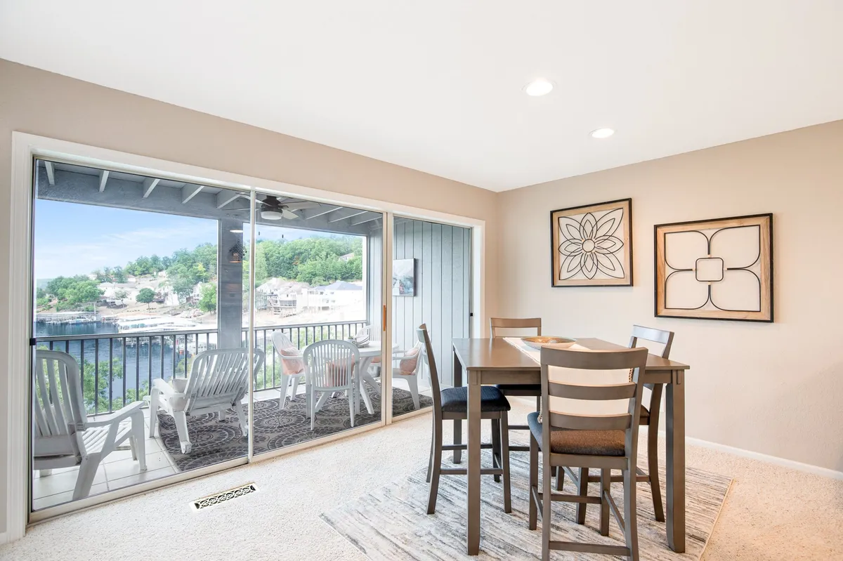 A dining table set near sliding glass doors that lead to an outdoor seating area on the balcony. The space is filled with natural light, features contemporary art and decor, and offers a direct sightline to the balcony and sparkling water beyond.
