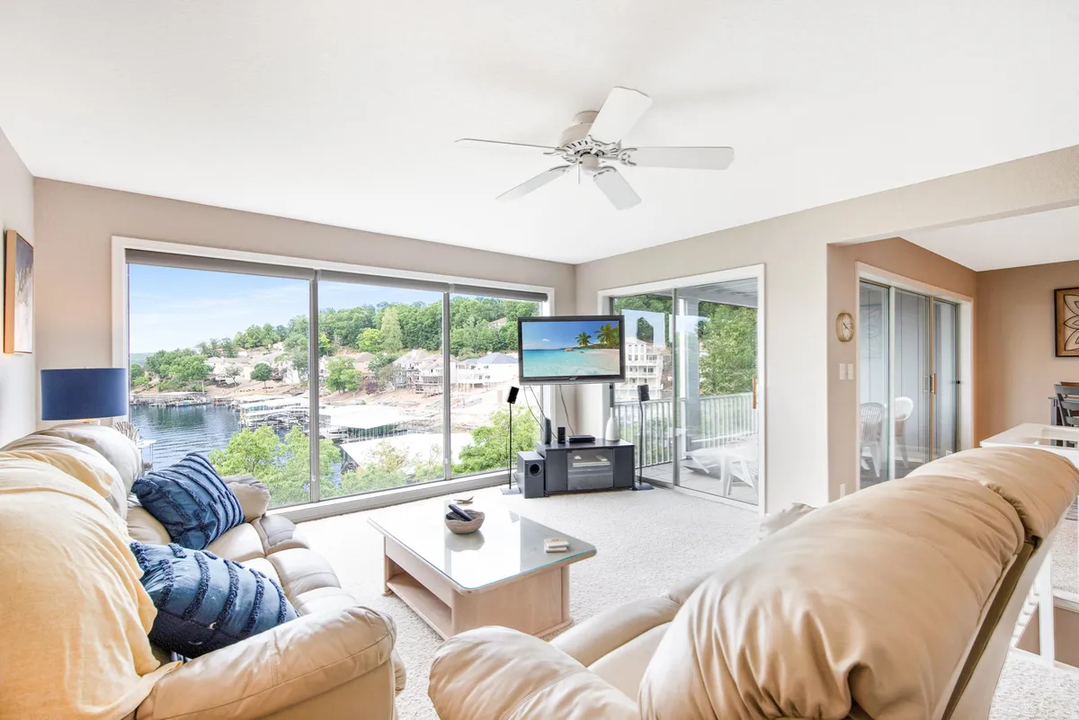 A cozy living room scene featuring large windows with unobstructed water views, comfortable couches, and a television. The room is arranged for group activities like movie nights or game sessions, with sunlight streaming in and the lake providing a serene backdrop.