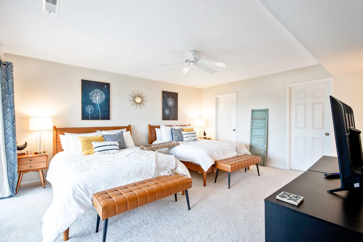 A serene bedroom featuring two beds adorned with decorative pillows, tasteful artwork on the walls, a wooden bench at the foot of the beds, and a bedside lamp. Soft natural light filters into the room, creating a calm and inviting atmosphere ideal for rest and relaxation.
