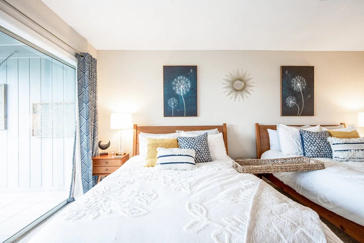 A comfortable bedroom with two beds, decorative pillows, subtle wall art, and a lamp. A sliding glass door opens to a balcony, inviting in sunlight and offering an indoor-outdoor flow. The setting is relaxed and flexible for families or multiple guests.