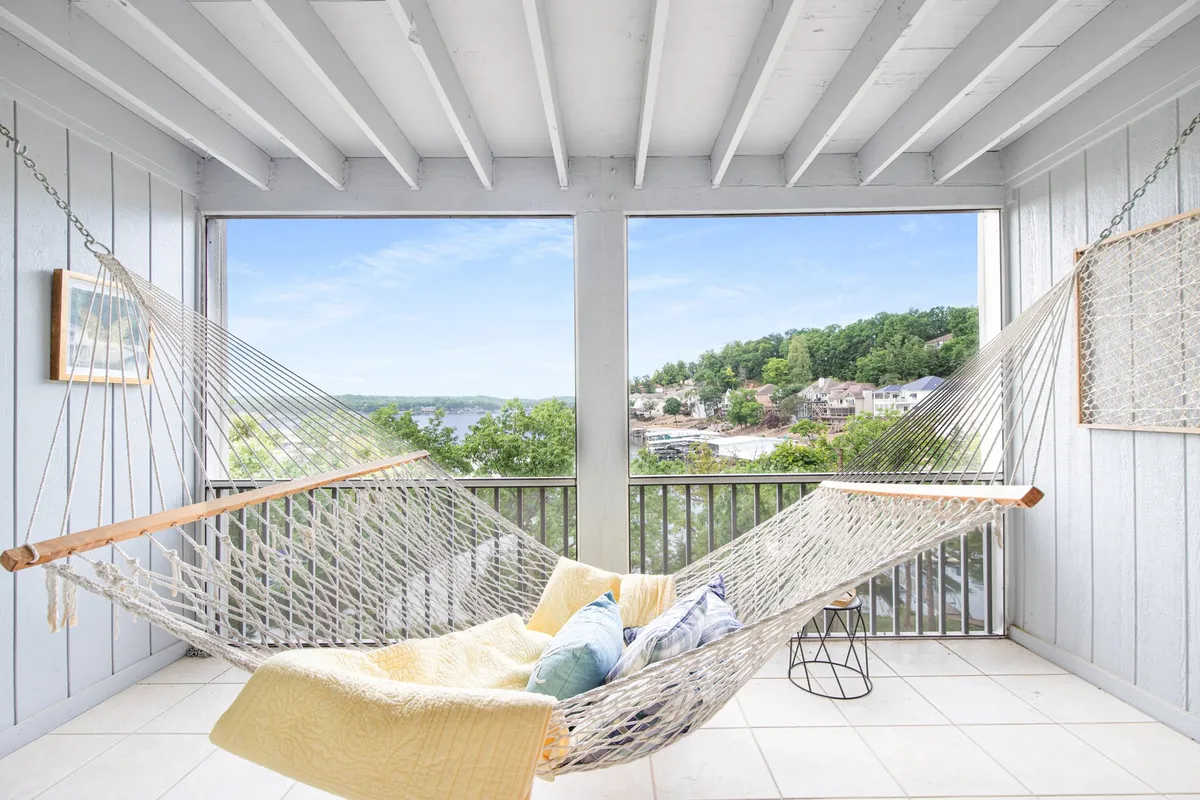 The image showcases a bright, inviting living space with a cozy hammock positioned near expansive windows that frame serene lake views. Sunlight filters through, casting a warm glow on the decorative wall hanging and plush throw, creating an atmosphere perfect for relaxing, reading, or simply soaking in the tranquil scenery.