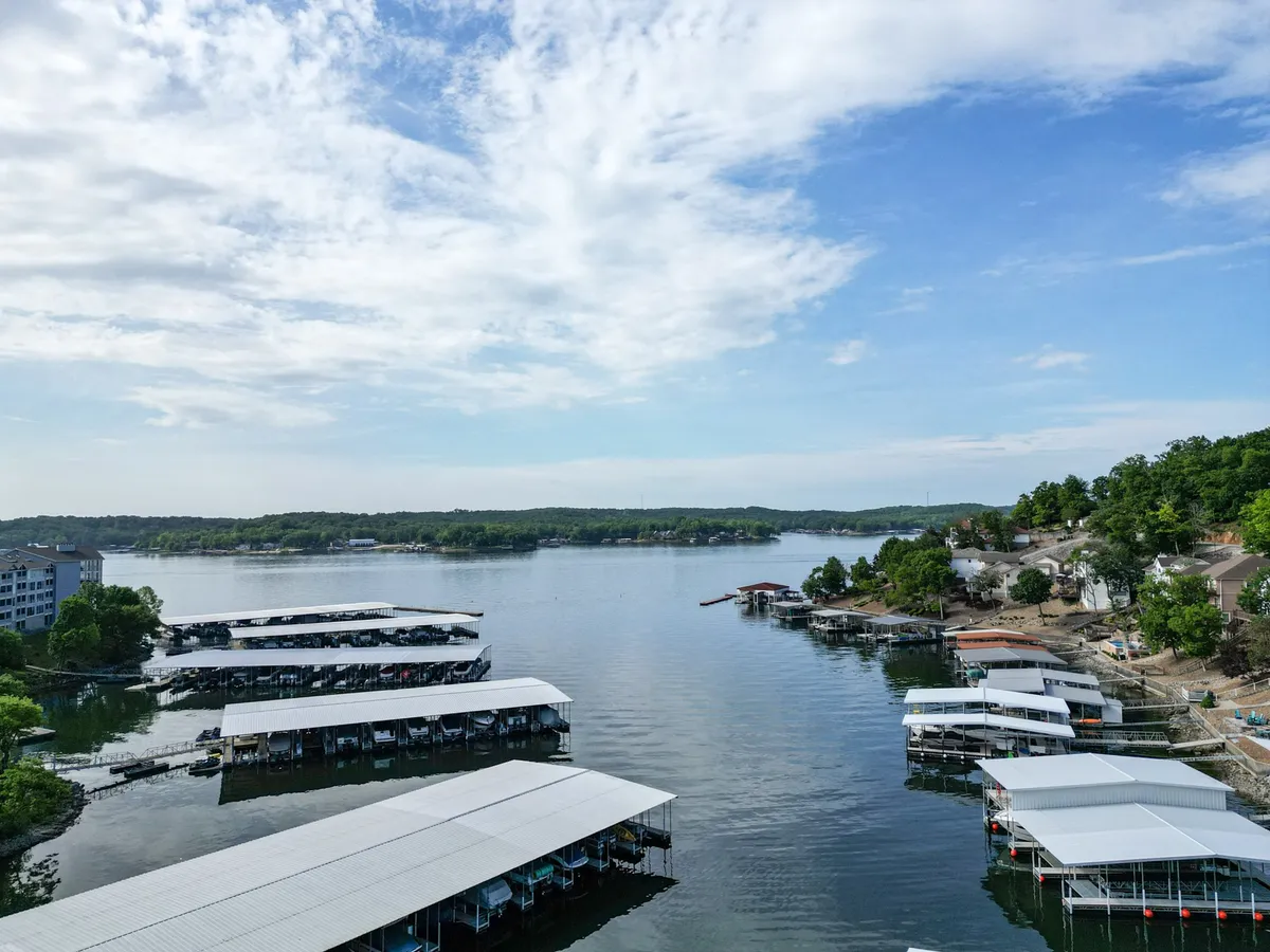 A panoramic view across calm lake waters, with several marinas, boats, and the surrounding greenery visible. The sky reflects beautifully on the water's surface, evoking a sense of relaxation and possibility for boating, fishing, or exploring nature trails.
