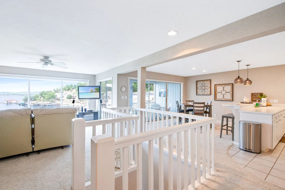 A spacious, open-plan living and dining area with large windows framing stunning lake views. Comfortable seating, a dining table, and a kitchen bar area create a cohesive, inviting social space. Natural light pours in, enhancing the airy atmosphere and blurring the boundary between indoors and the lake outside.