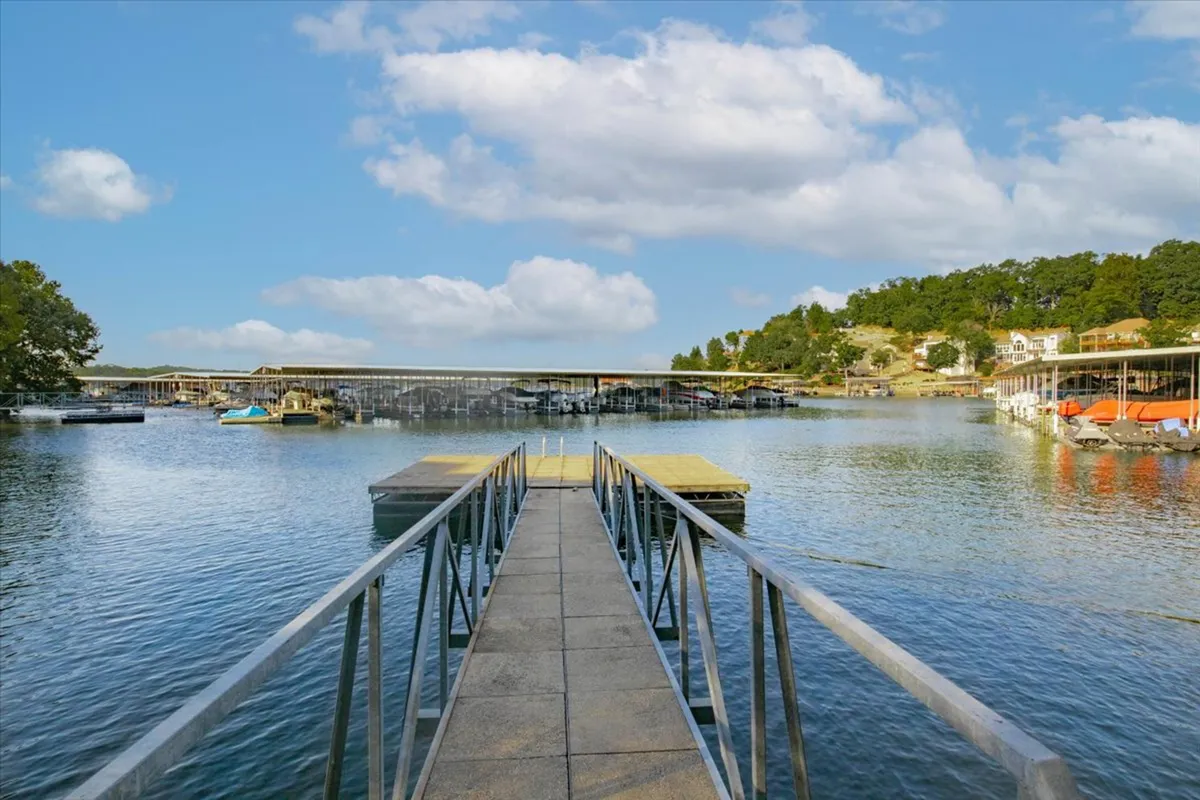 A tranquil lakeside dock extends into the water, with boats moored nearby and a boathouse visible. Hillside homes overlook the scene, and the area feels peaceful yet ready for activity. People can be seen enjoying the dock, suggesting opportunities for launching boats, fishing, or simply relaxing by the water.