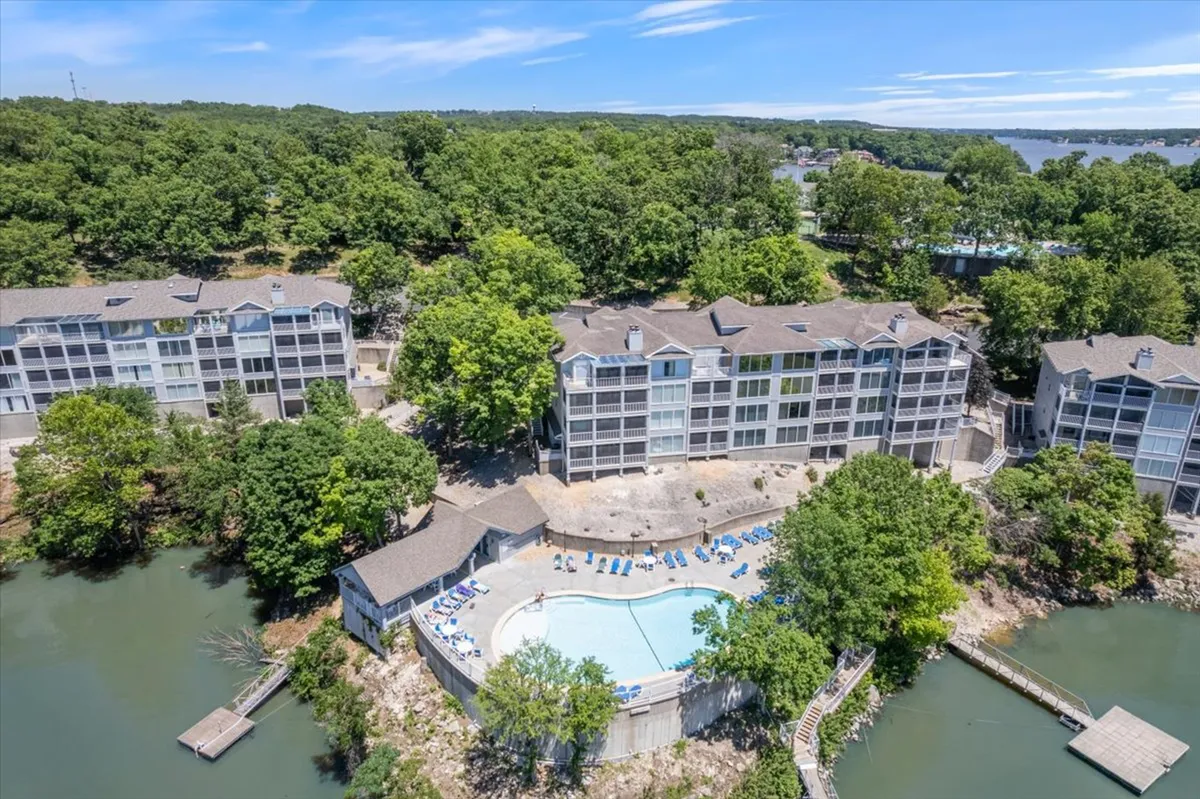 A welcoming pool area set against a backdrop of lush greenery and waterfront buildings. The space is arranged for both fun and relaxation, with lounge chairs and room for children to play. The water and surrounding condominiums are visible, hinting at a vibrant community atmosphere close to the lake.