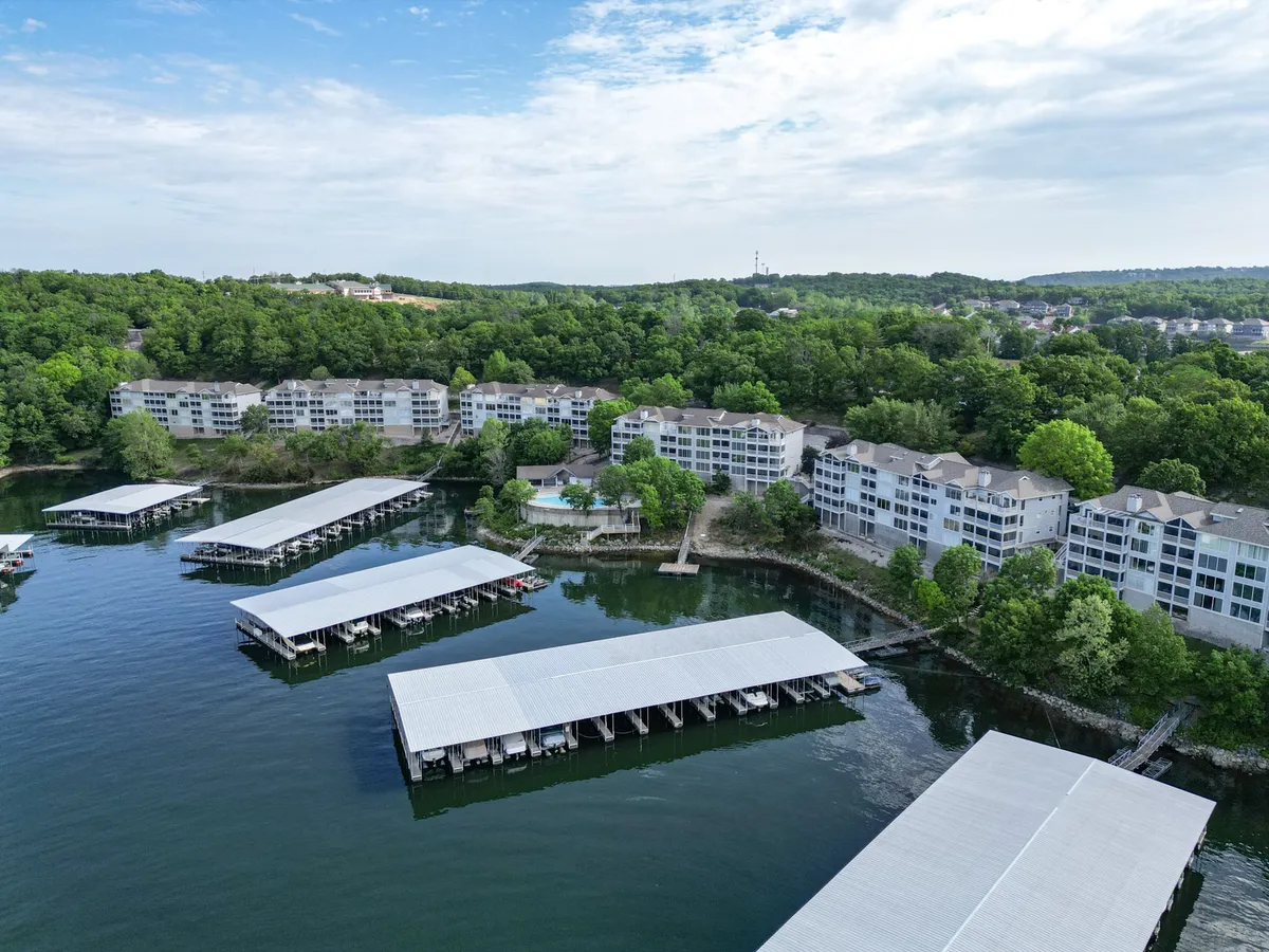 A tranquil lakeside scene featuring the shimmering lake, several boathouses along the shoreline, lush greenery, and nearby residential buildings. The pool area is visible in the foreground, offering options for swimming and relaxation. The scene is bathed in soft light, suggesting either morning coffee or a peaceful sunset.