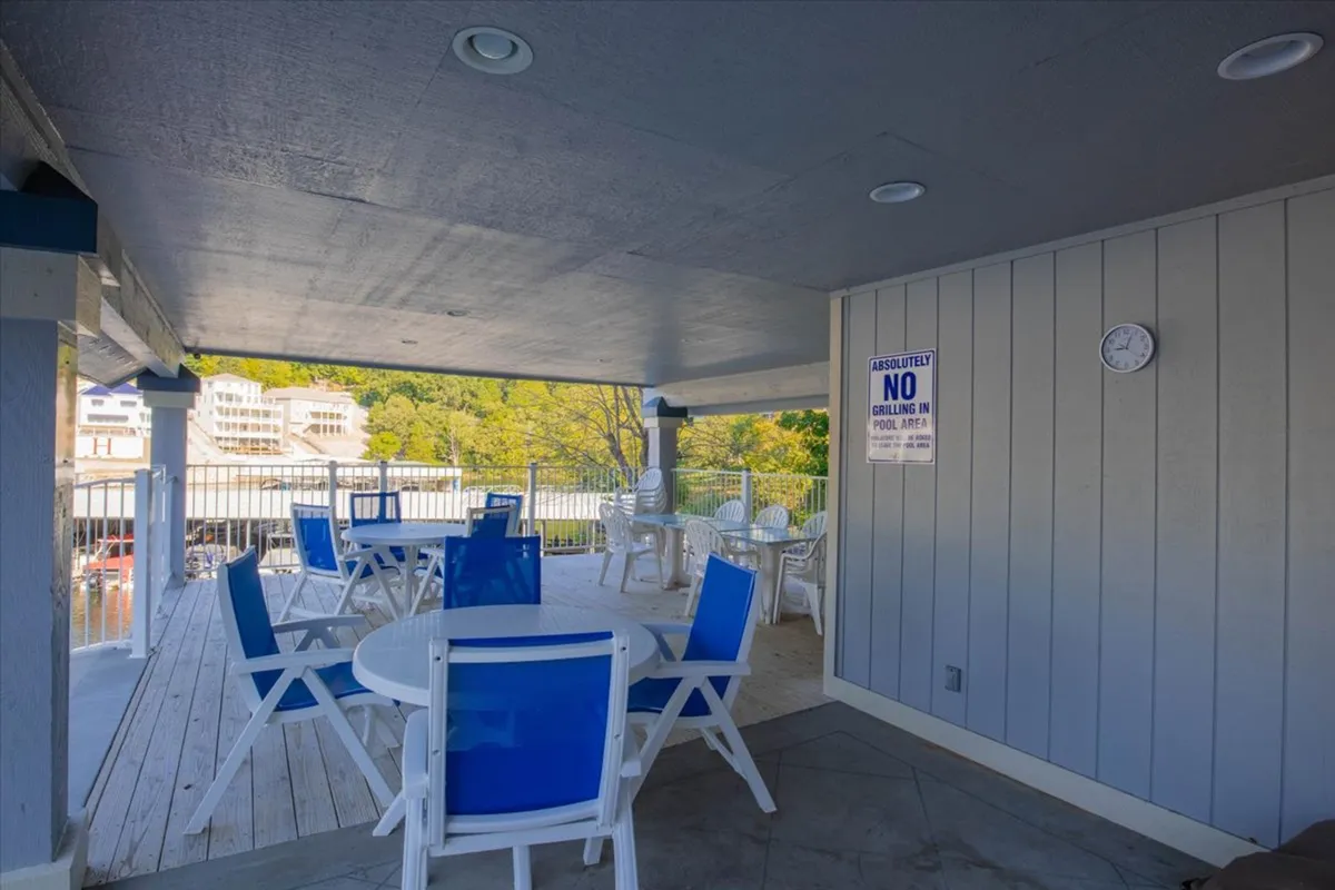 A resort-style pool area is surrounded by white tables and blue chairs, set against the warm glow of sunset. Groups of people are seen relaxing, socializing, and enjoying the atmosphere, with visible signage enhancing the community feel.